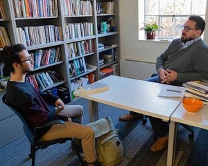 Male professor meeting with male student in book-lined faculty office.