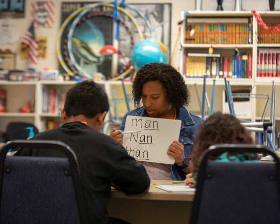 Holding a flash card, a Black woman helps two children with a language lesson in an elementary classroom.