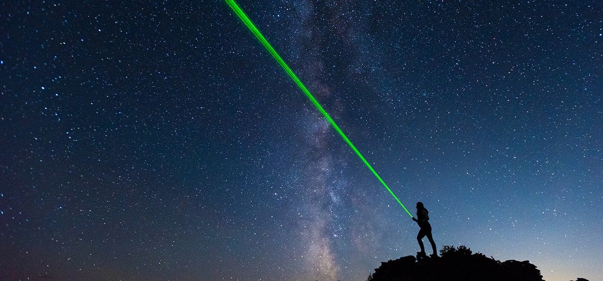 Silhouette of researcher on hilltop, shining a green beam into starry nighttime sky.