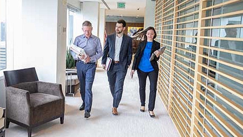 Three lawyers in conversation, carrying files and walking down hallway in an office building.