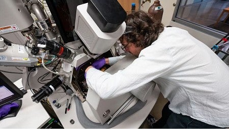 A white male wearing a white lab coat and purple neoprene gloves stoops to work on high-tech lab equipment.